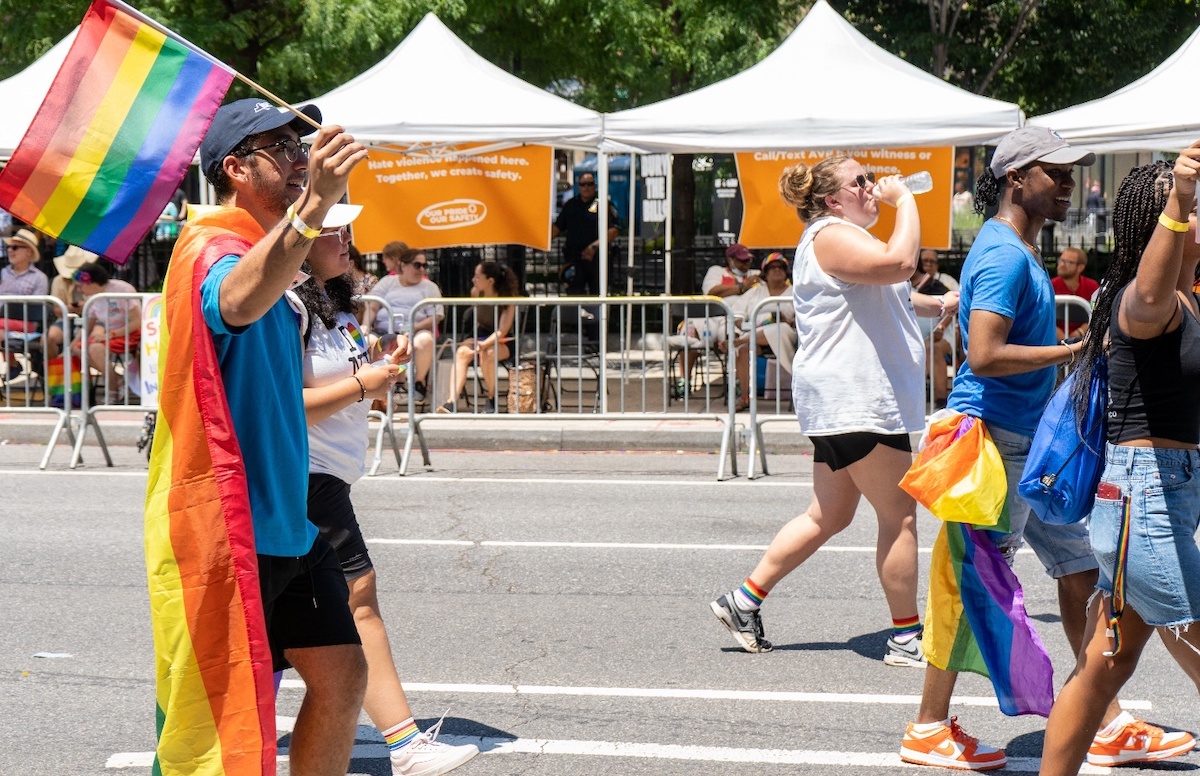 People marching in NYC Pride carrying rainbow flags in front of orange AVP signs.